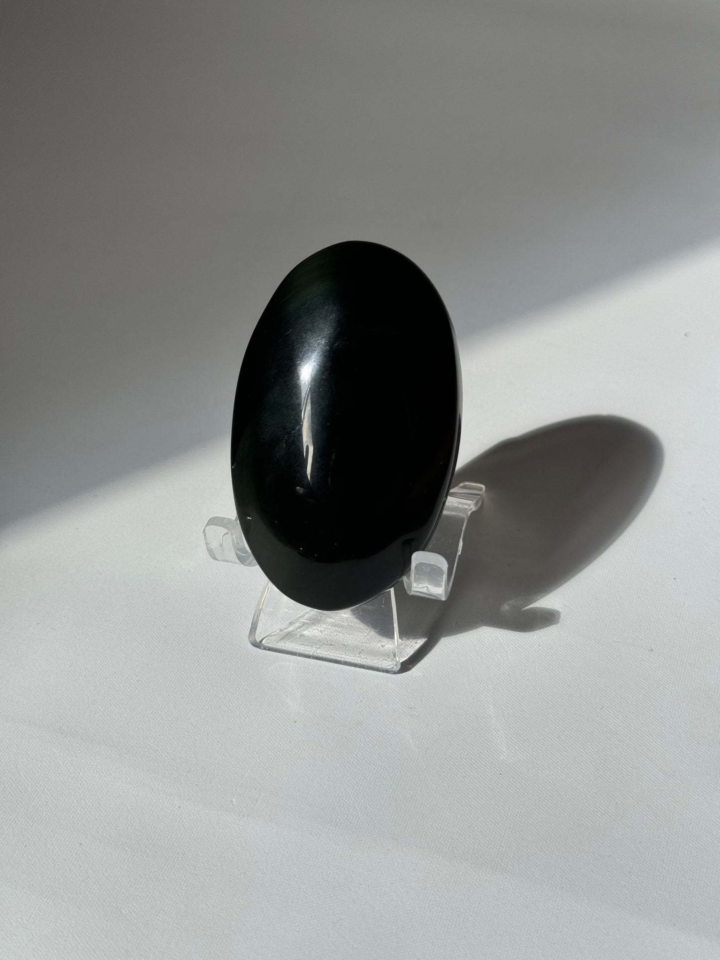 Rainbow Obsidian Crystal Palm Stone on a transparent stand, revealing hints of green and gold hues under natural light.