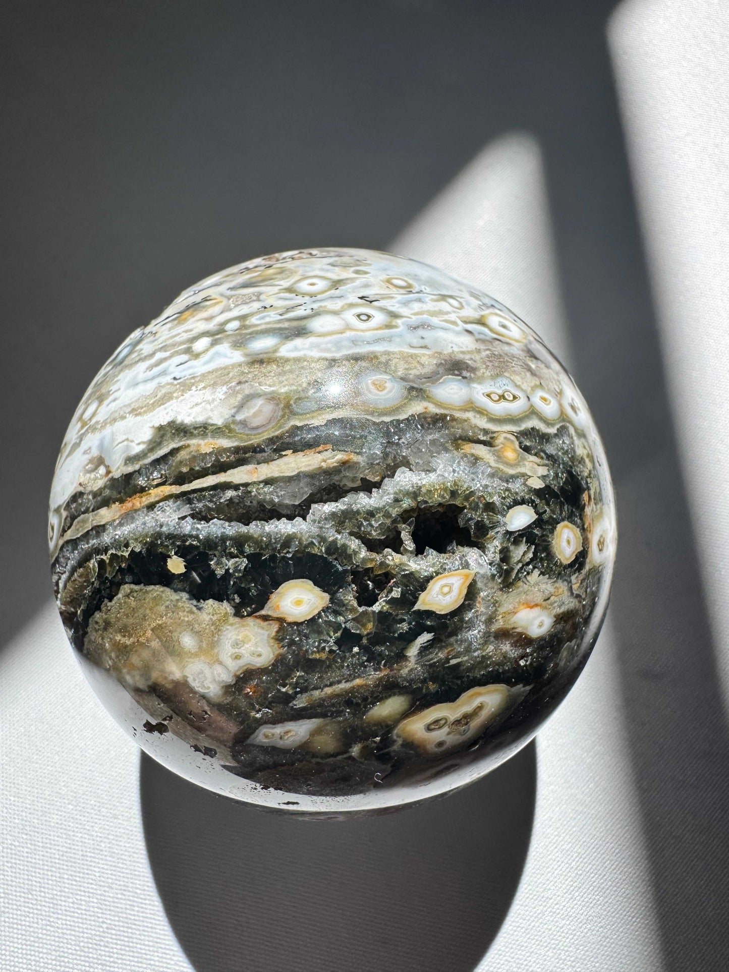 Orbicular Ocean Jasper Sphere #4 showcasing green, white, and beige orb patterns with natural crystal cavities under direct light. Ideal for gemstone collectors | Bespoke Crystals Auckland NZ