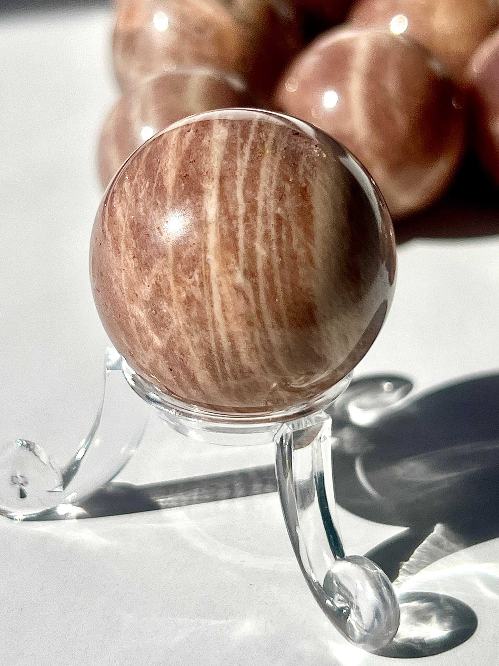 Peach Moonstone Crystal Mini Sphere on a clear stand, showcasing natural striations and warm tones, perfect for enhancing perception and inner growth.