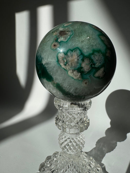 Green Flower Agate Crystal Sphere #1 displayed on a faceted glass stand, showcasing soft green hues and floral-like mineral patterns.