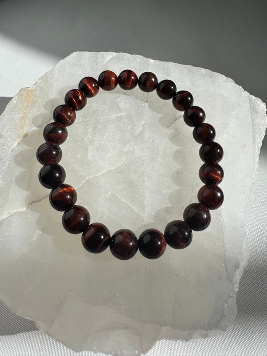Red Tigers Eye Crystal Bracelet with polished round beads on a white quartz slab, showcasing deep reddish-brown hues and silky luster.