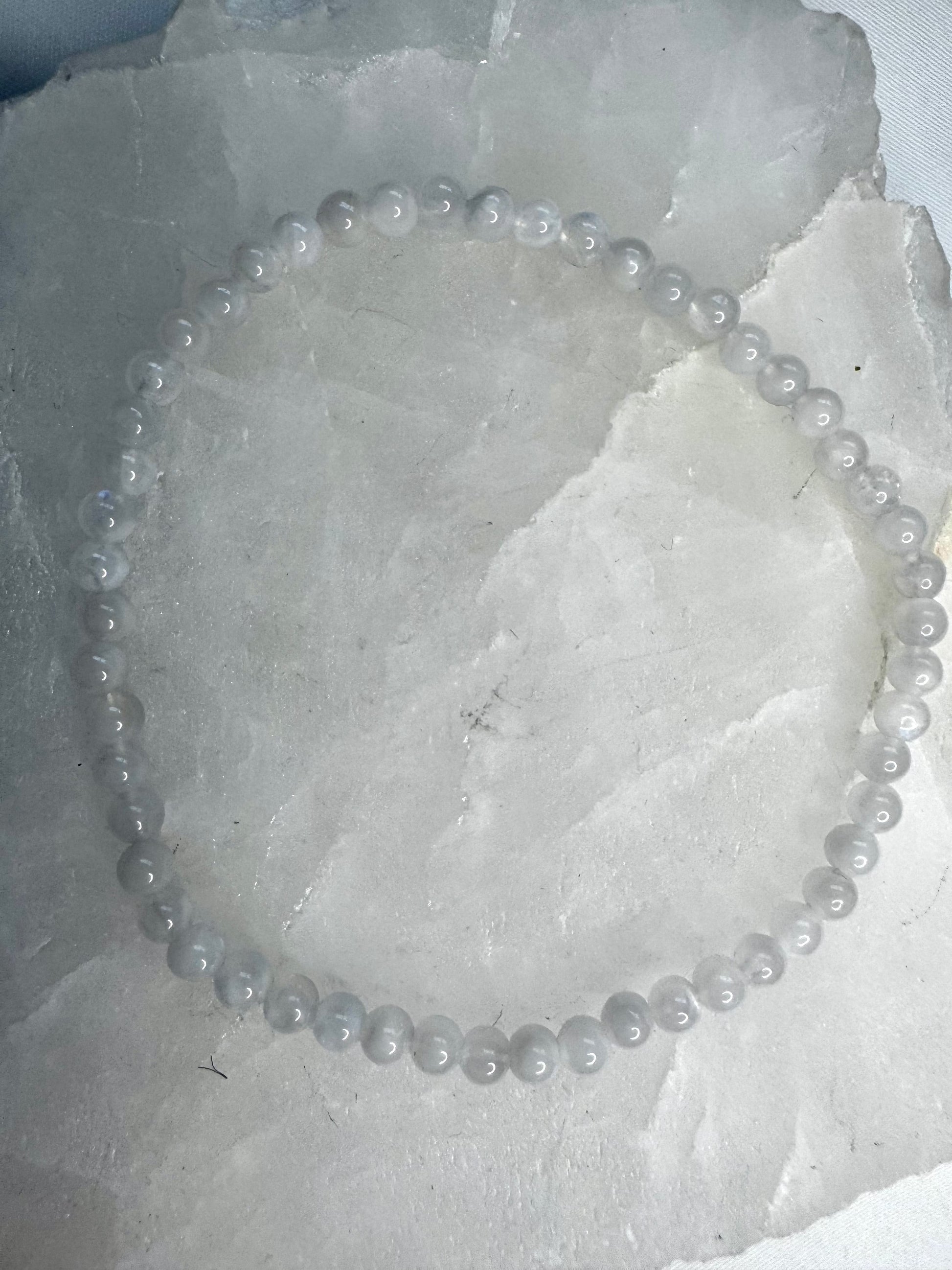 Single strand Moonstone Crystal Bracelet 4mm with polished round beads, laid flat on a white crystal background. Promotes intuition and spiritual growth.