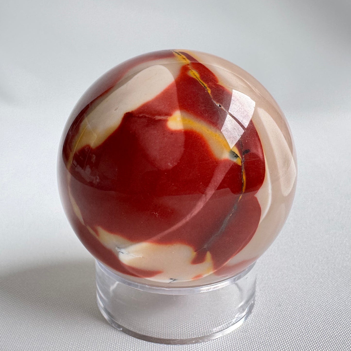 Mookite Crystal Sphere | Bespoke Crystals featuring deep red, cream, and yellow tones on a clear stand. Ideal for grounding and creative energy.