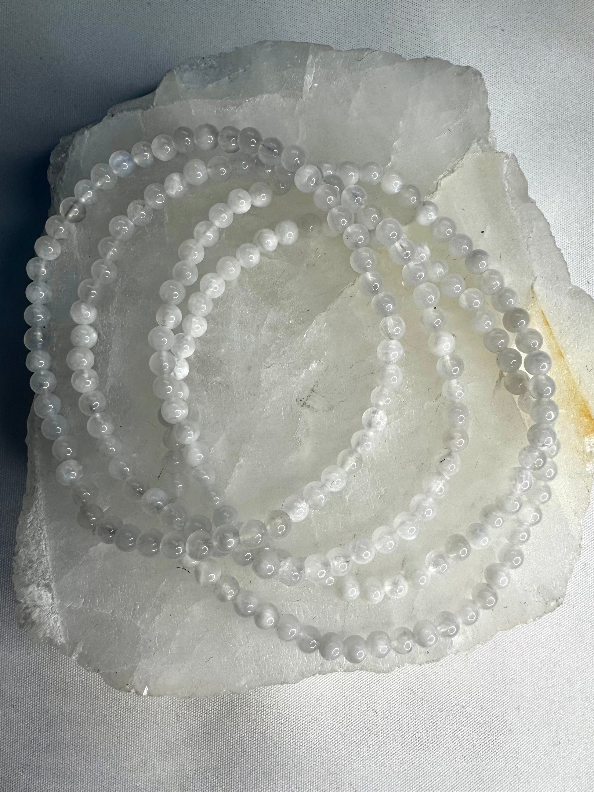 Moonstone Crystal Bracelet 4mm featuring luminous white beads with adularescence, displayed coiled on a raw quartz slab. Ideal for emotional balance and creativity.