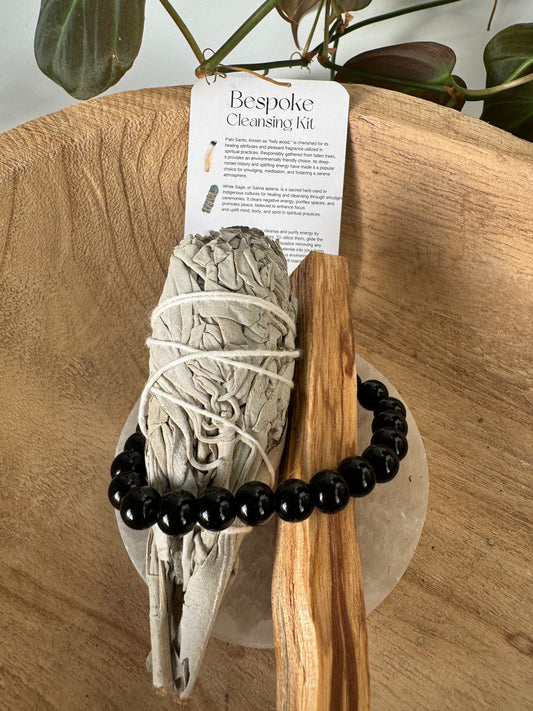 Bespoke Crystal Cleansing Kit featuring sage bundle, Palo Santo stick, black crystal bracelet, and instruction card on wooden tray. Ideal for energy cleansing rituals.