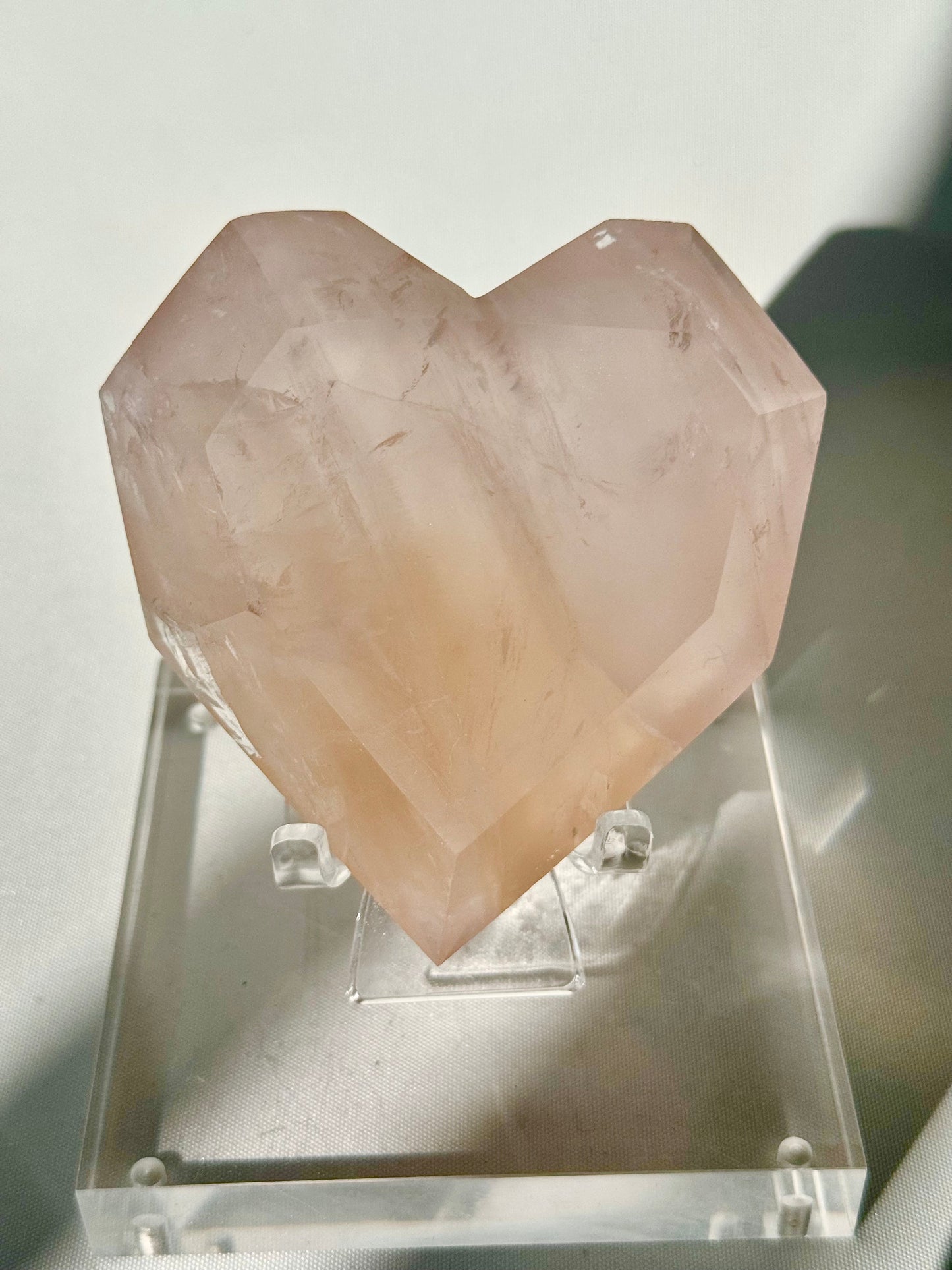 Faceted Rose Quartz Crystal Heart with soft pink hues and natural inclusions, displayed on a clear stand under direct light.