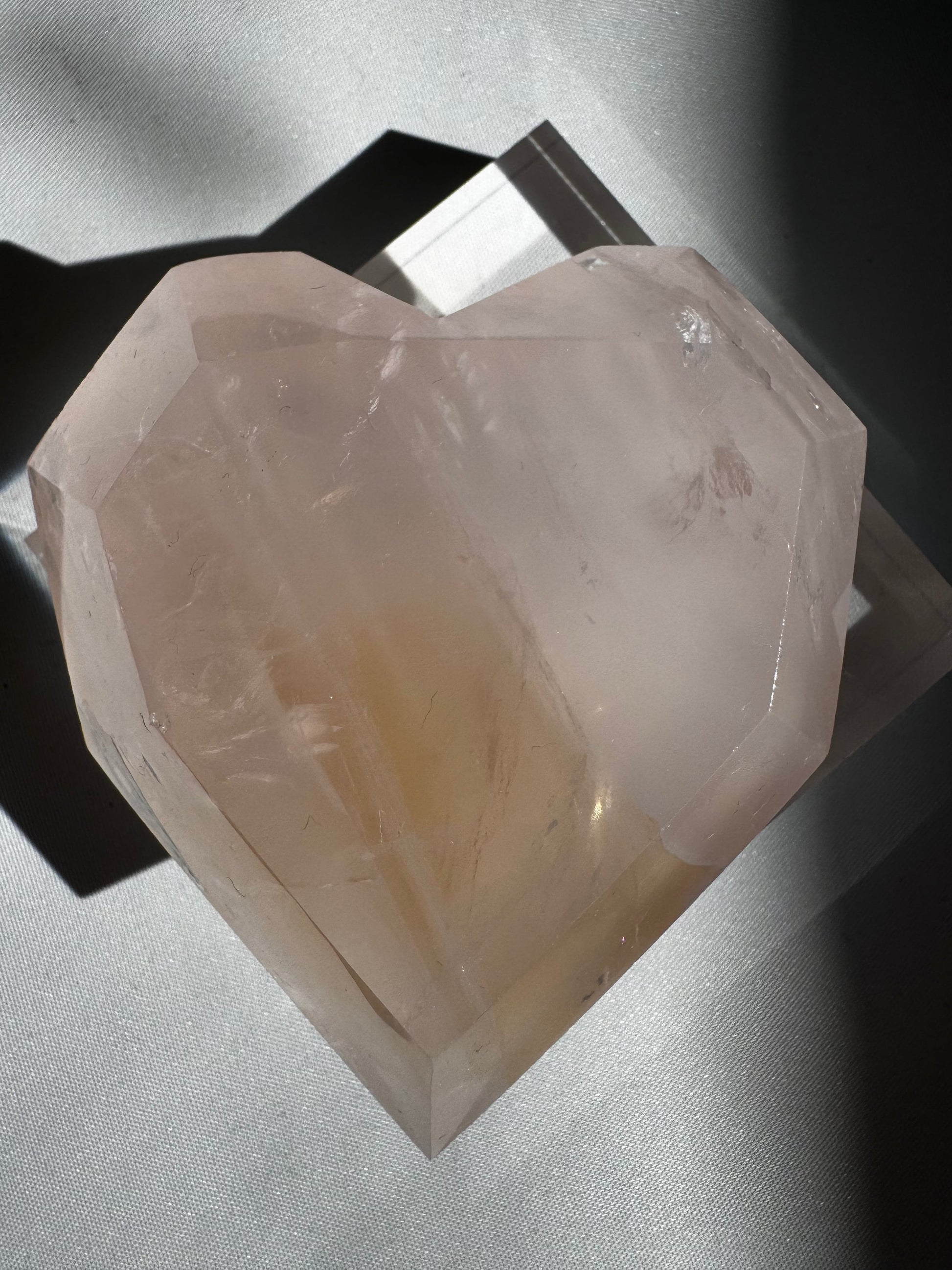 High-quality Rose Quartz Crystal Heart with faceted edges and calming pink tones, partially shadowed on a display base.