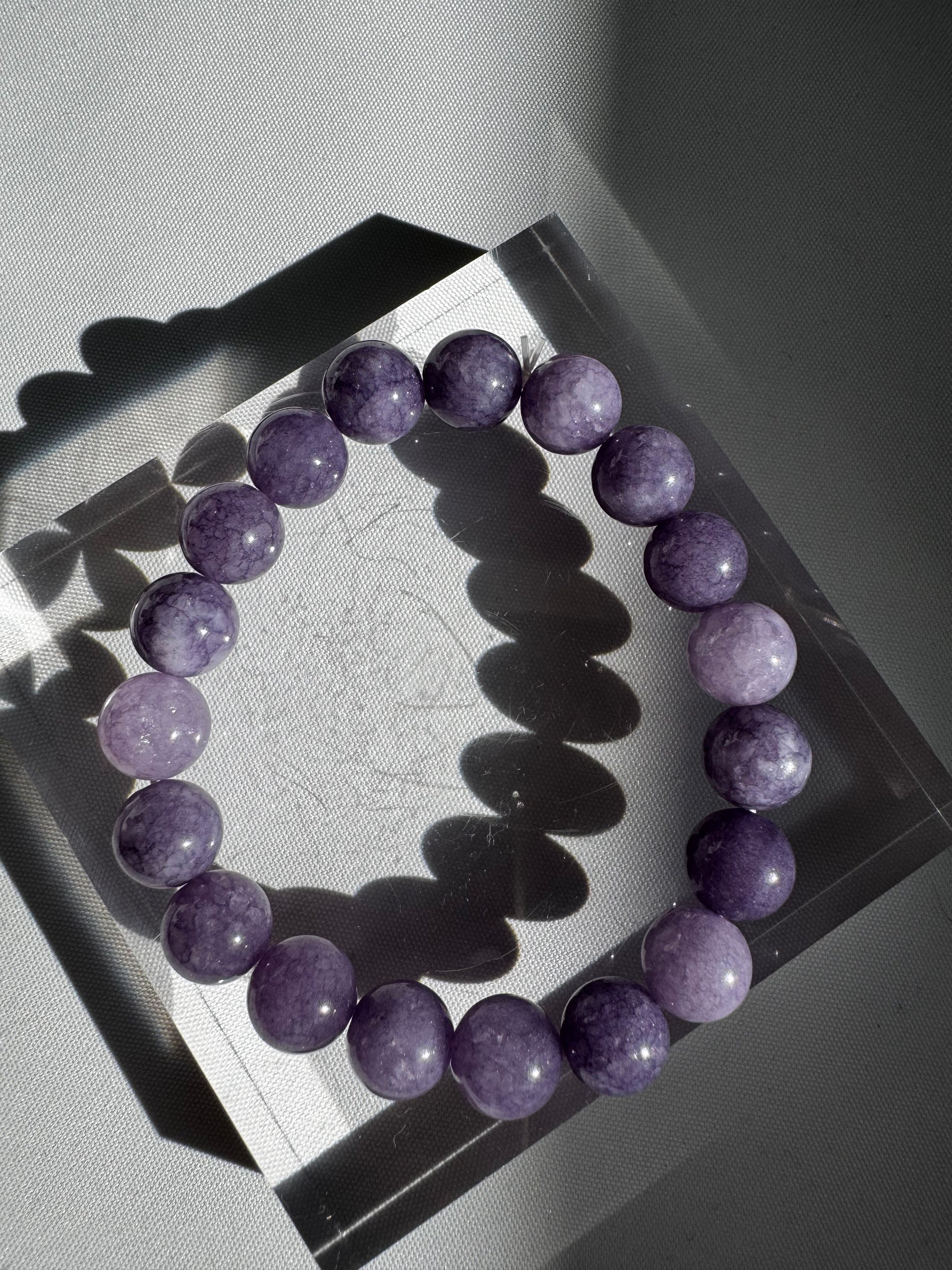 Single 10mm Lavender Jade Crystal Bracelet displayed on a clear block, showcasing its vibrant purple tones and smooth, glossy finish.