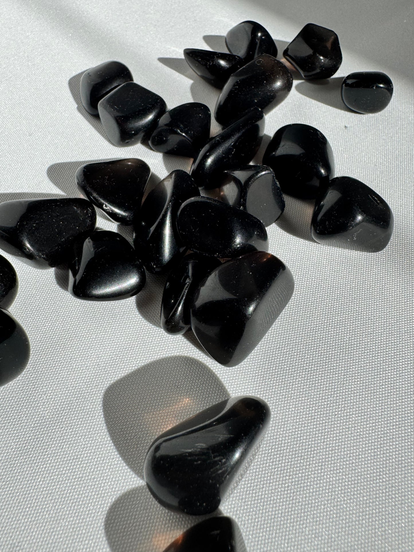 Glossy Apache Tears tumble stones for healing and protection, displayed on white fabric under natural light. Ideal for emotional release and grounding.