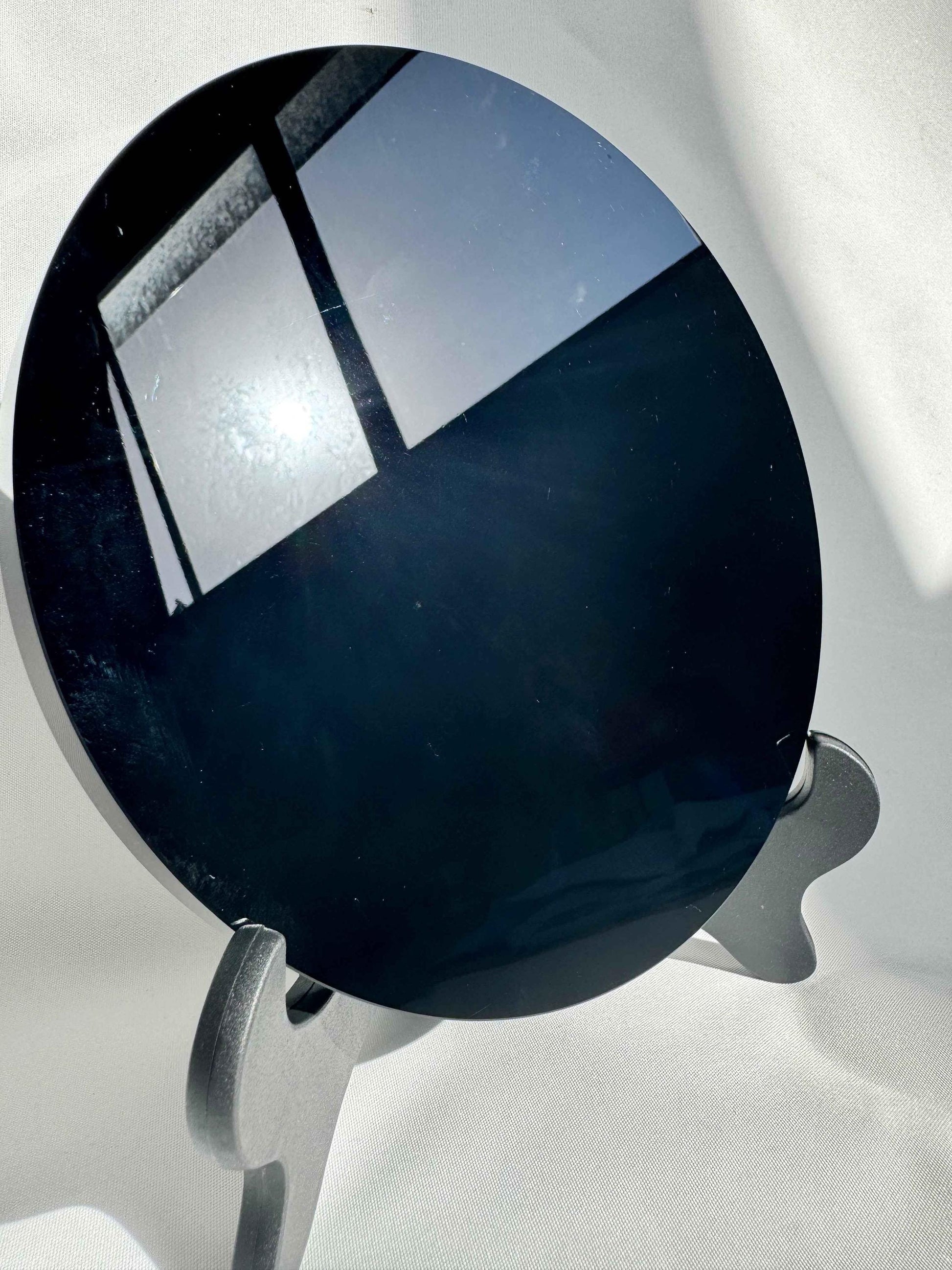 Polished Black Obsidian Crystal Mirror reflecting sunlight, displayed on a black stand. Ideal for metaphysical use