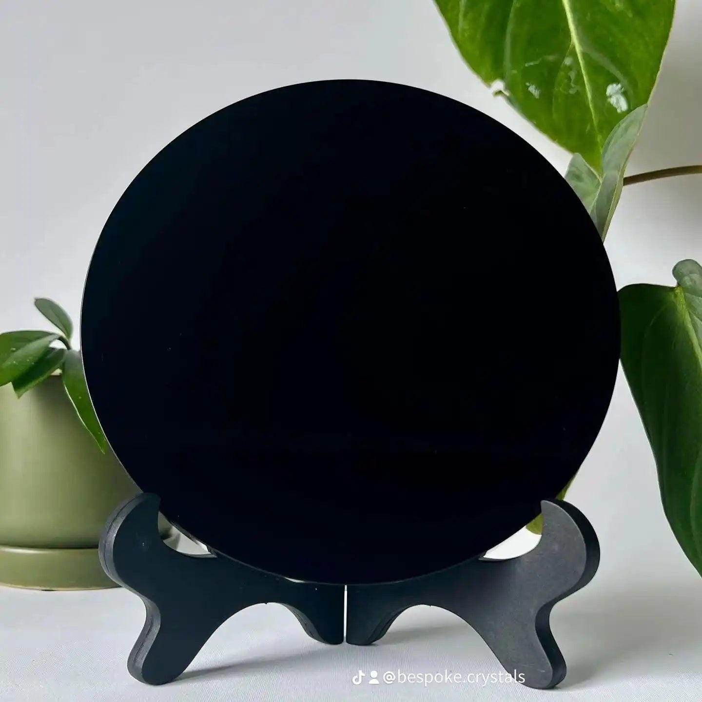 Black Obsidian Crystal Mirror on black stand, surrounded by green leaves. A collectible from Bespoke Crystals for grounding and protection.